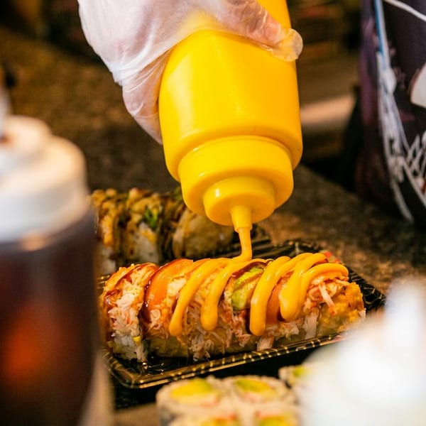 A gloved hand squeezes yellow sauce from a bottle onto a sushi roll in a black tray.