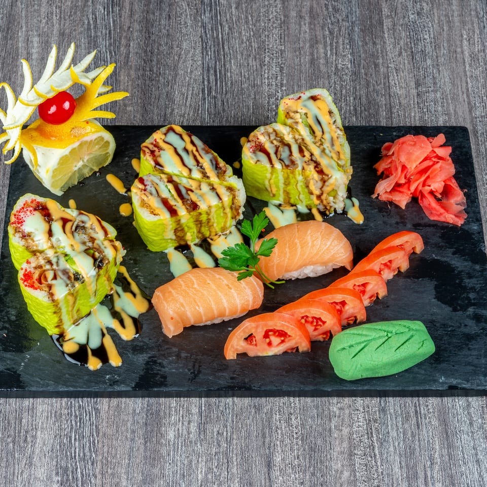 Sushi platter with green rolls, salmon nigiri, tomato slices, ginger, wasabi, and lemon garnish.