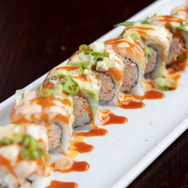 Sushi rolls topped with creamy and spicy sauces, avocado, and sliced green onions on a white rectangular plate.