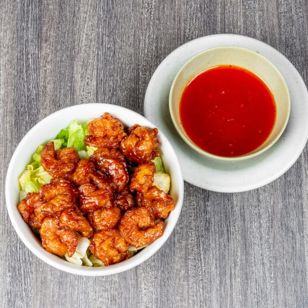 Sweet and sour shrimp with lettuce in a white bowl, next to a small bowl of red sauce.