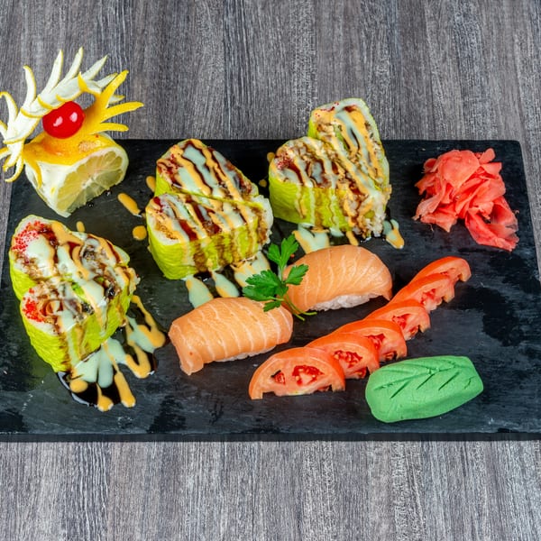 Sushi platter with green rolls, salmon nigiri, tomato slices, pickled ginger, and wasabi on a black board.
