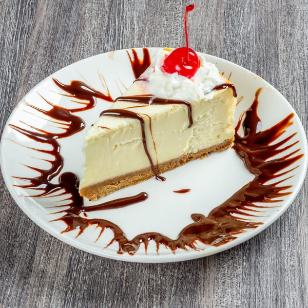 Slice of cheesecake with chocolate drizzle, whipped cream, and a maraschino cherry on a white plate.