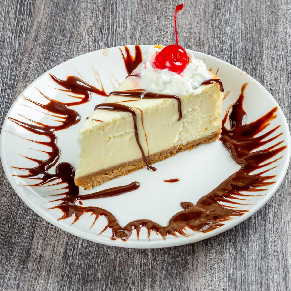 Slice of cheesecake with chocolate drizzle, whipped cream, and a maraschino cherry on a white plate.